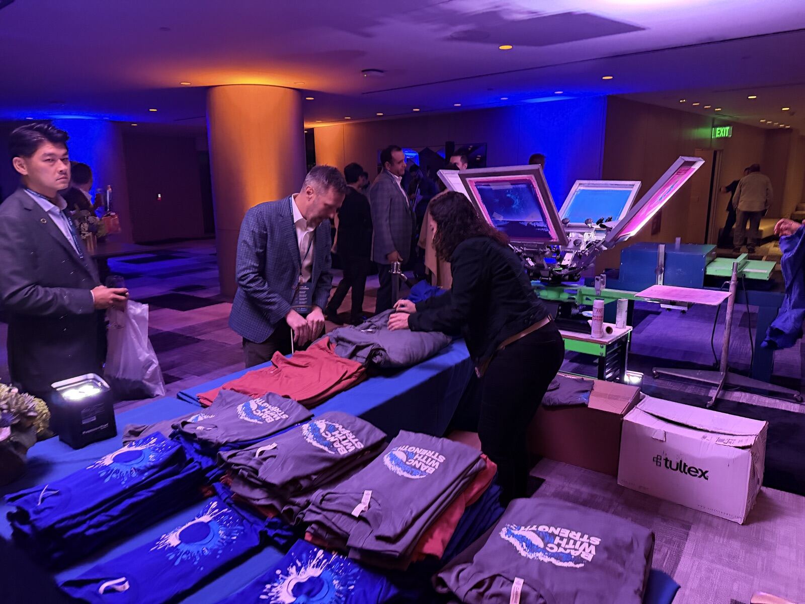 Corporate gala guests in blazers gathering around a garment personalization table under blue uplighting