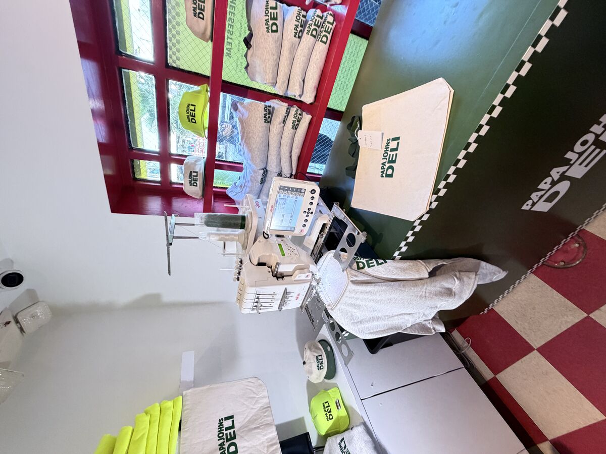 Commercial embroidery machine running at a branded deli pop-up with neon caps and canvas totes staged around it